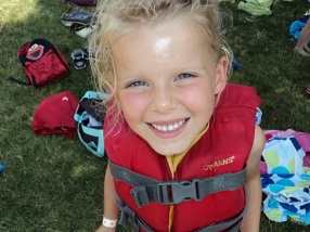 a little girl in a life jacket stands and smiles upwards towards the camera